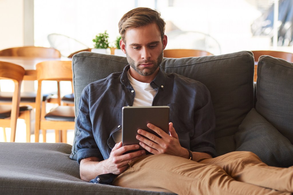 Man sitting on a gray couch using a tablet in a bright living room.
